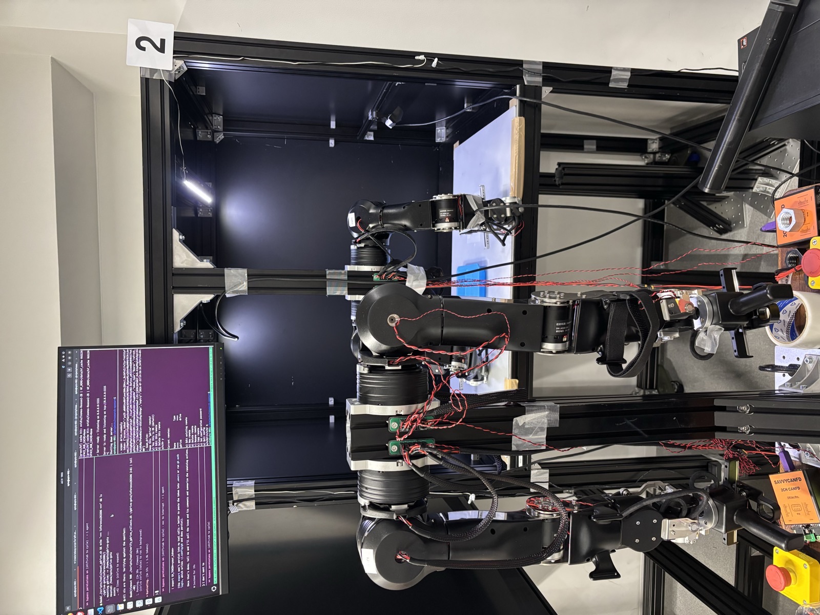 The recording cell: leader arms in the foreground, the bimanual OpenArm followers inside an aluminum-extrusion cell with the workspace board, tray, cameras and LED line lights; a monitor above shows the recorder terminal