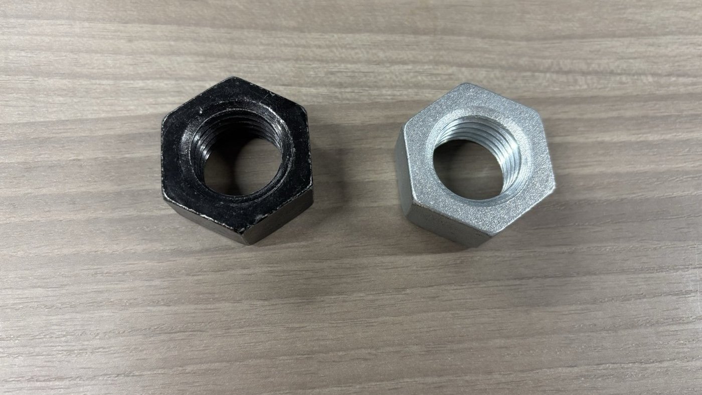 Two hex nuts on a wooden surface: a black one on the left and a bright silver one on the right